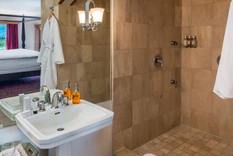 Large tiled shower with pedestal sink, white robe hanging and view of bedroom in the mirror