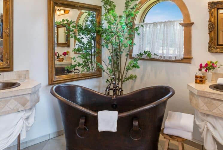 Unique copper soaking tub in the corner of a bathroom flanked by two sinks with gold framed mirrors