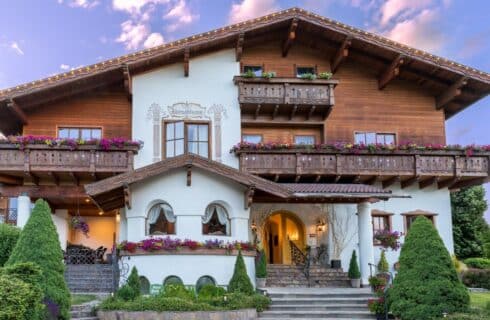 Front facade of a three story white and brown home in old word style wiith balconies lined in flowers, lush landscaping and arched window details