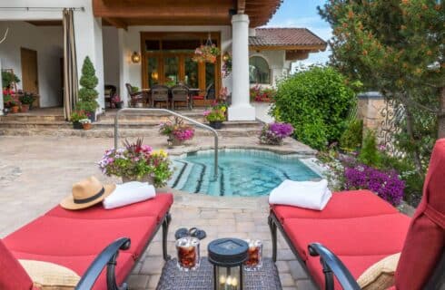 Spacious outdoor patio of a luxury two story home with hot tub, red lounging chairs, patio table and chairs and flower pots all over