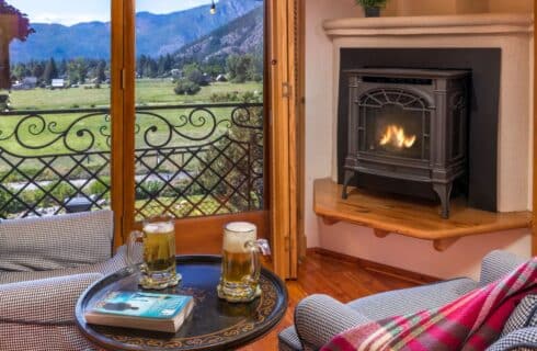 Cozy sitting area with two upholstered sitting chairs, table with two mugs of beer, small fireplace and large patio doors overlooking the mountains