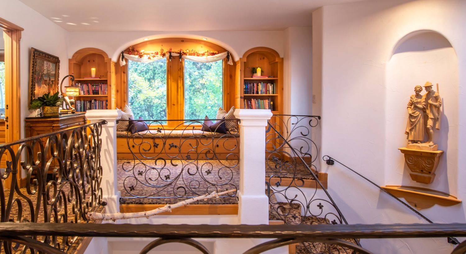 Elegant window seat flanked by two arched built in bookcases at the top of a landing with a wrought iron railing with white columns