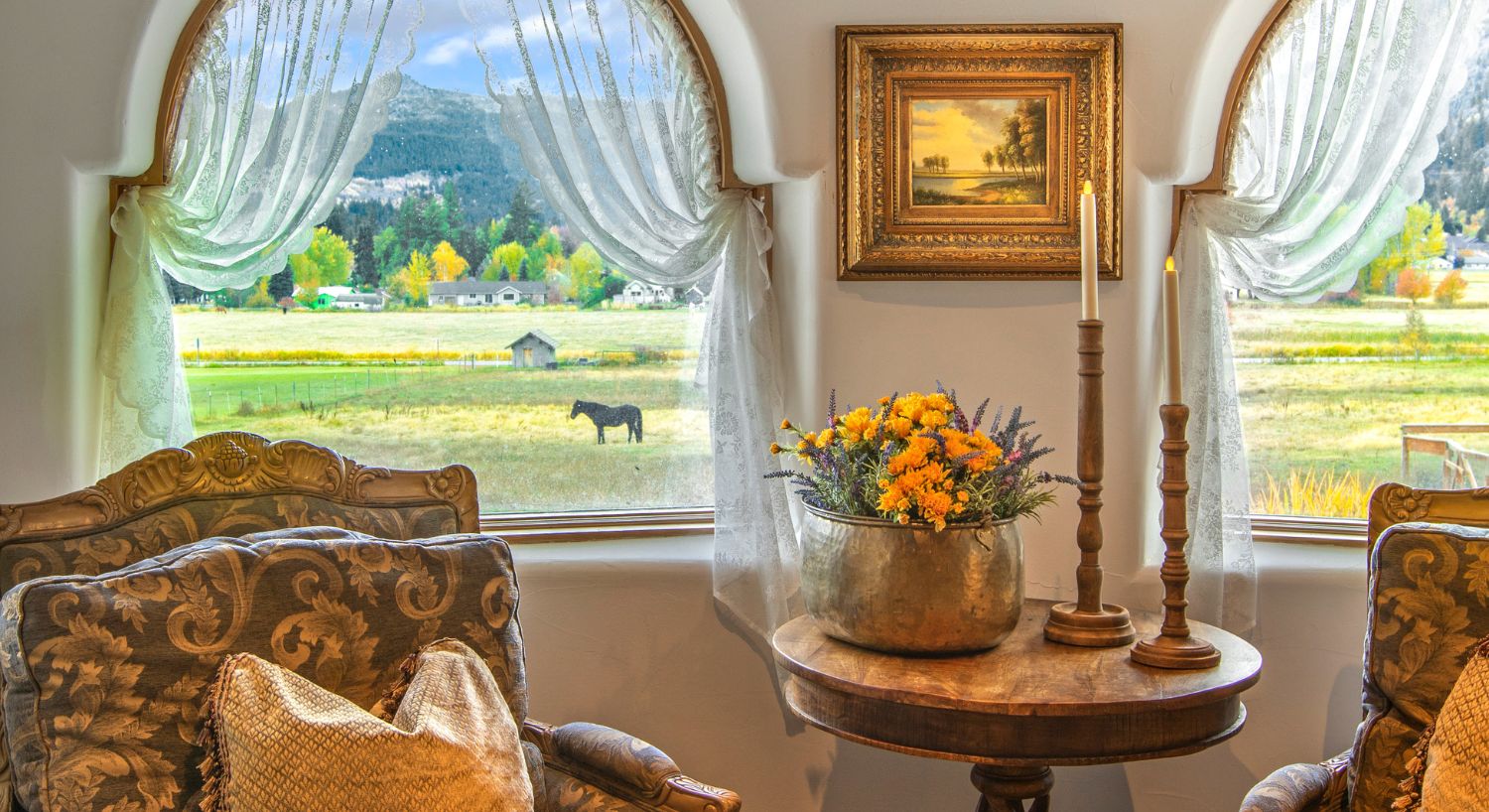 Sitting area with two plush decorative chairs and table in front of large arched windows overlooking a valley and mountains