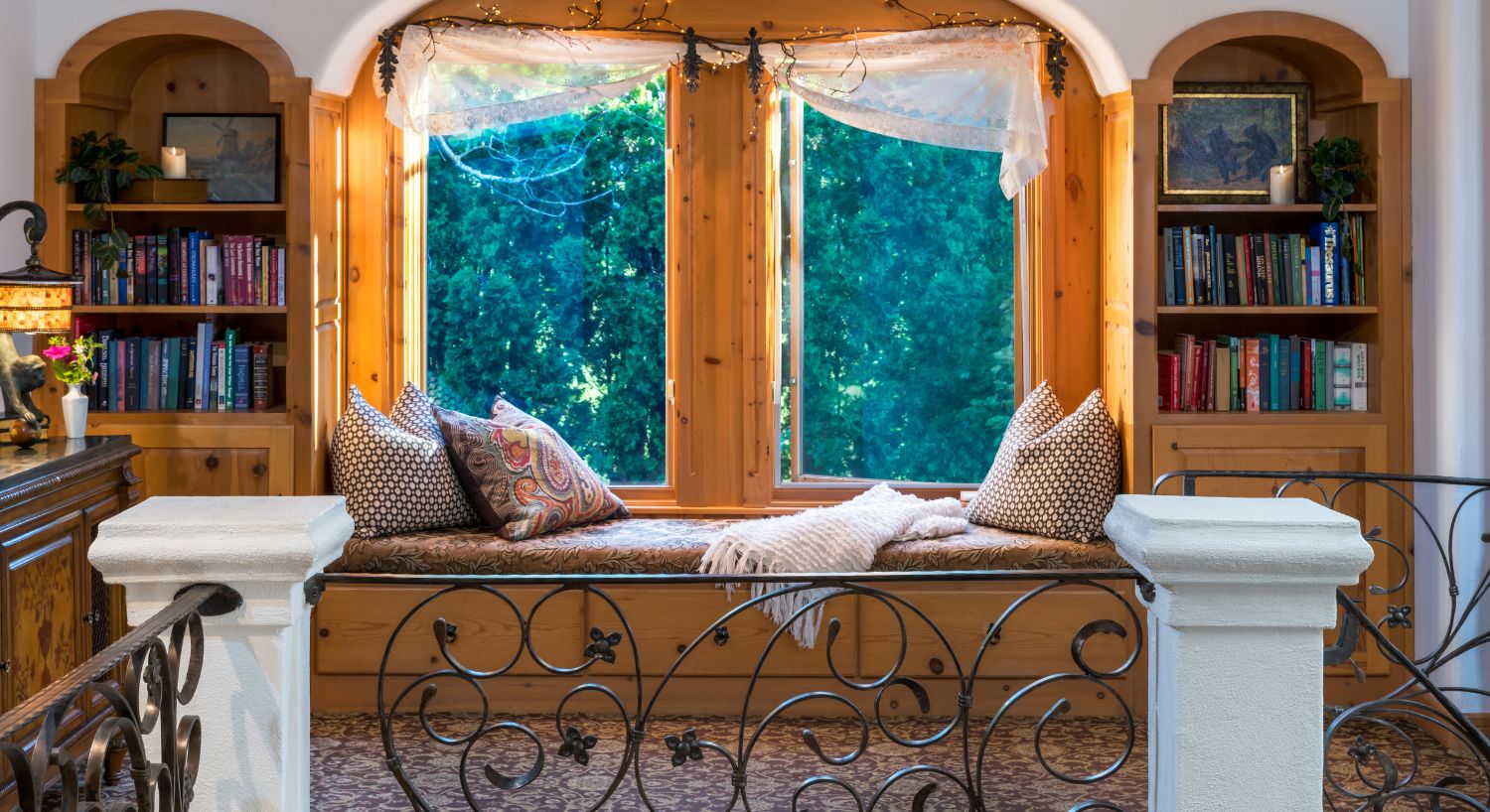 A beautiful window seat flanked by two arched bookcases, in front of a wrought iron railing with white columns