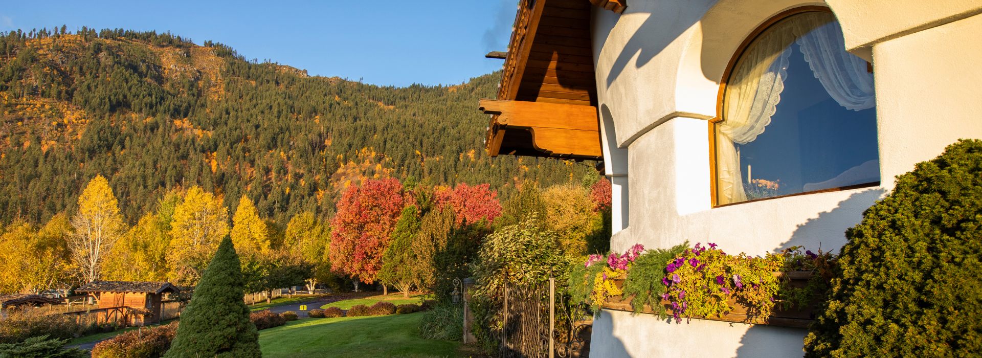 Side view of a unique white and brown home with arched window details and flower boxes, at the base of a mountain with fall colored trees