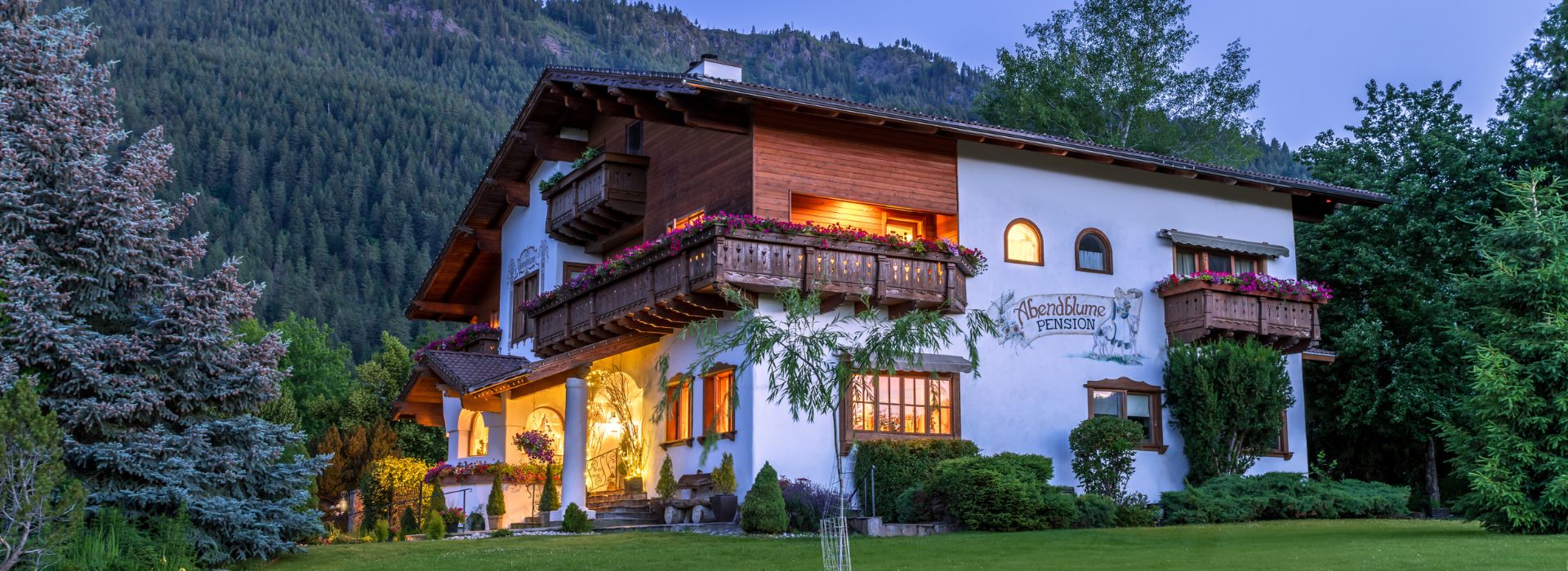 Large three story white and brown home in Bavarian style architecture with golden lights at dusk at the base of a tree covered mountain