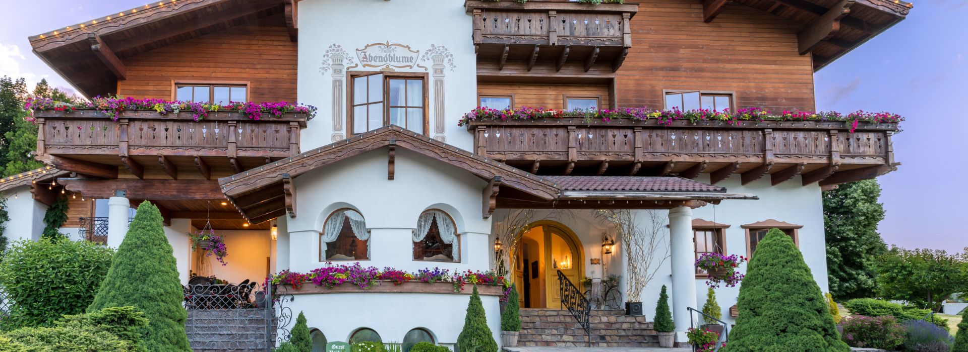 Front facade of a three story white and brown home in old word style wiith balconies lined in flowers, lush landscaping and arched window details