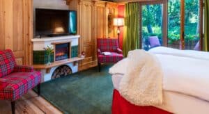 Cozy bedroom with king bed in white linens, fireplace and TV flanked by two plaid sitting chairs, door open to a patio