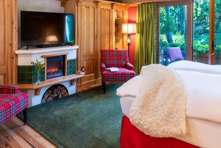 Cozy bedroom with king bed in white linens, fireplace and TV flanked by two plaid sitting chairs, door open to a patio