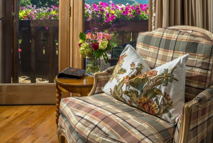 A plush sitting chair in plaid fabric with a decorative pillow in front of a patio door with view of colorful flowers