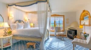 Bright and elegant bedroom with canopy bed with white drapery, fireplace, sitting chairs and large window