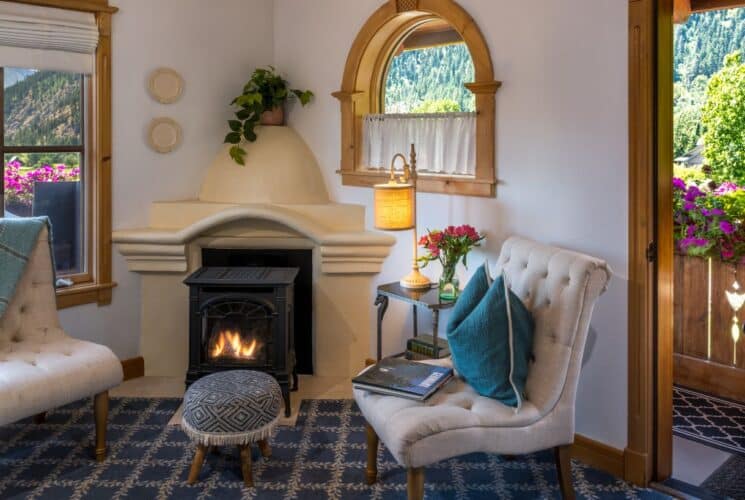 Sitting area of a bedroom with two white chairs and footstool in front of a decorative fireplace, large windows and patio door open to a balcony