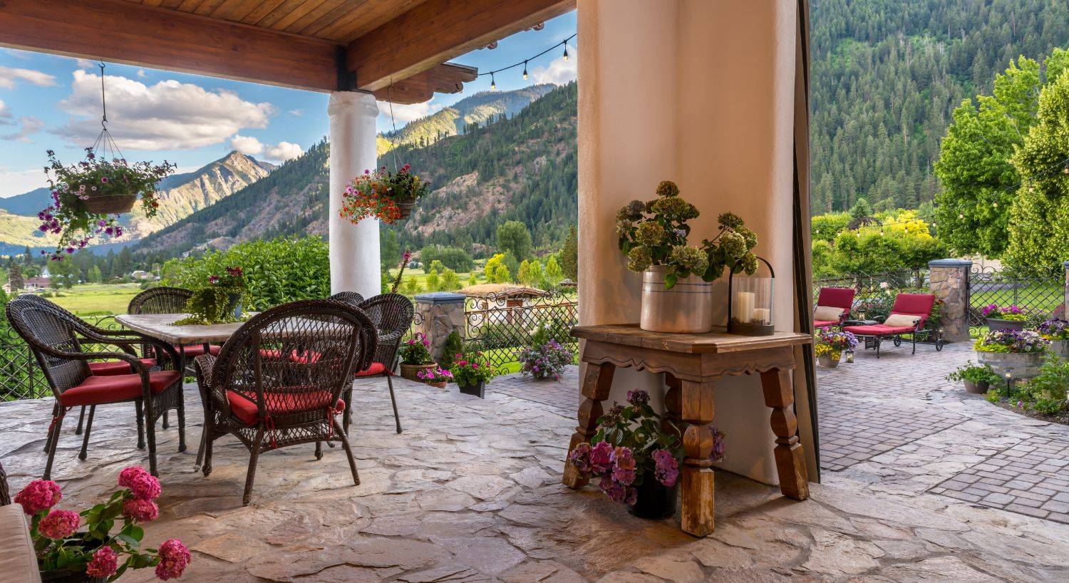 Expansive outdoor patio with decorative stone, wicker furniture, flower pots and string lights overlooking a vast mountain range