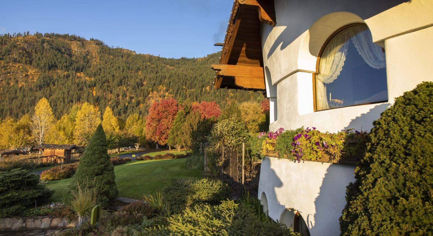 Side of a white and brown home with arched windows and flower boxes against a mountainside of fall colored trees