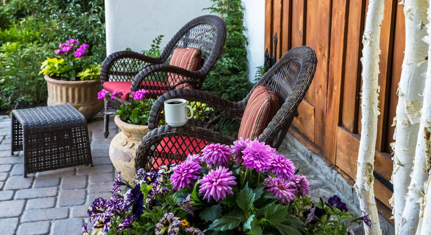 Two wicker sitting chairs with red and striped cushions on a brick patio by wood doors and flower pots