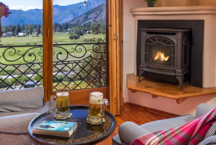 Two upholstered sitting chairs and a table with mugs of beer in front of a small fireplace, by a large patio door overlooking a mountain range